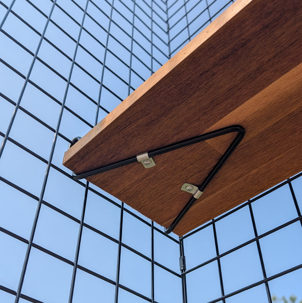 A Shelf Bracket with Hardware (no wood) is mounted on a black wire grid wall, supporting a wooden shelf, with a clear blue sky visible in the background.