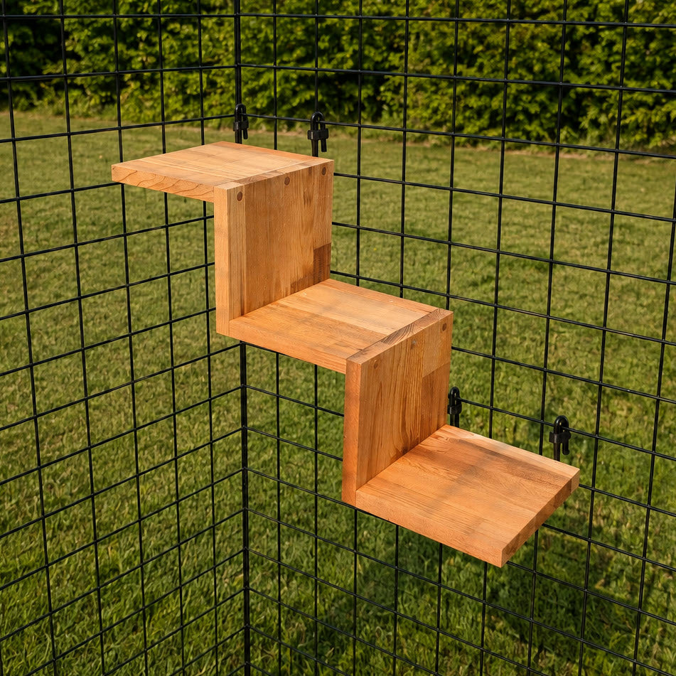 The Staircase: Cedar Three Step Kit For Catios is attached to a black wire grid fence in a grassy outdoor area with green bushes in the background, creating a playful feature for any cat enclosure.